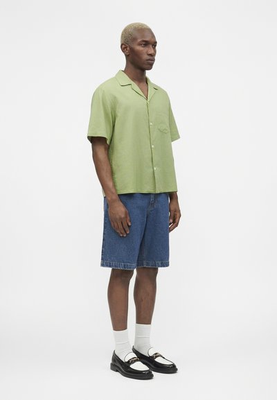 Άνδρας με κοντά ξανθά μαλλιά, ντυμένος με ανοιχτά πράσινο κοντομάνικο πουκάμισο με κουμπιά, τζιν σορτς μέχρι το γόνατο, λευκές κάλτσες και ασπρόμαυρα μοκασίνια.