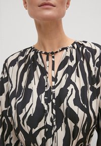 Schwarz- und cremefarbige gemusterte Bluse mit rundem Halsausschnitt, versehen mit einem seitlichen Schlüssellochdetail und weichem, leichtem Stoff. Faltendetails sind vorhanden.