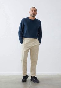 Námořnická košile s dlouhým rukávem v kombinaci s béžovými kalhotami, které mají přiléhavý střih. Černé tenisky doplňují outfit na neutrálním pozadí.