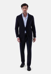 Mann, der einen strukturierten dunkelblauen Blazer, ein hellblaues Hemd, dunkle Hosen und schwarze Slipper trägt, vor einem schlichten Hintergrund steht.