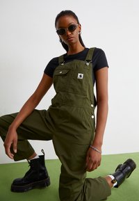 Olive green cotton overalls with adjustable straps, side buttons, and front pockets. Paired with black platform boots and sunglasses.