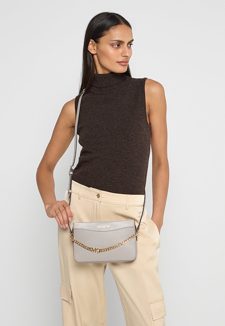 Grijze crossbody tas met kettingriem, gladde leren textuur, rechthoekige vorm, logo detail aan de voorkant, gecombineerd met beige broeken en een bruine top.