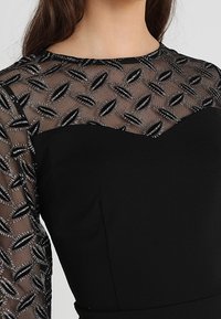 Svart klänning med en genomskinlig broderad meshöverdela som har texturerade ovala mönster, kompletterad av en åtsittande nedre del i fast svart.