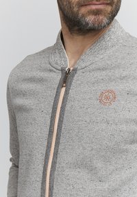 Graue Jacke mit Reißverschluss und geripptem Kragen, mit einer subtilen Textur, einem orangefarbenen Logo und einem kontrastierenden Reißverschlussdetail an der Vorderseite.