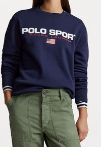 Mörkblå sweatshirt med "POLO SPORT" och "RALPH LAUREN" i fetvitt, med en liten amerikansk flagga och randiga ärmslut.