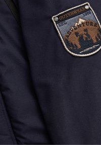 Tissu bleu foncé avec une pièce rectangulaire cousue représentant une scène de montagne, des arbres, et le texte "OUTERWEAR", "EST. 1986" et "ADVENTURE".