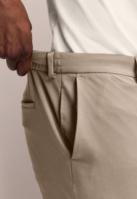 Pantalones beige con una textura suave, delantero con pliegues, pasadores para cinturón y un bolsillo lateral; una mano sosteniendo la cinturilla demuestra el tamaño.