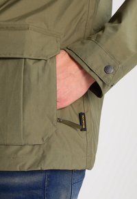 Main insérée dans la poche d'une veste vert olive avec étiquette Texapore, à côté du poignet avec bouton-pression en métal, portée avec un jean bleu.