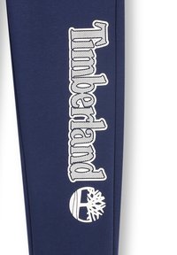 Marineblauwe stof met witte "Timberland" logo en icoon. Het materiaal lijkt glad met een lichte glans. Het ontwerp heeft een verticale plaatsing.