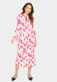 Rochie lungă, lejeră, cu plisuri verticale, având un model tie-dye roz pe un fundal alb și mâneci lungi. Balerini negri cu vârf ascuțit.