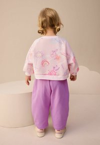 Jasnoróżowa bluza z kolorowymi wzorami, w tym kwiatami i tęczami, w połączeniu z luźnymi fioletowymi spodniami. Miękka tkanina, casualowy design.