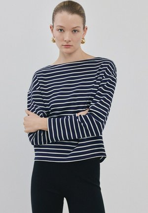 Jonge vrouw met haar naar achteren gebonden, gouden creolen, een marineblauw-wit gestreept shirt met lange mouwen en een zwarte broek, tegen een effen achtergrond.