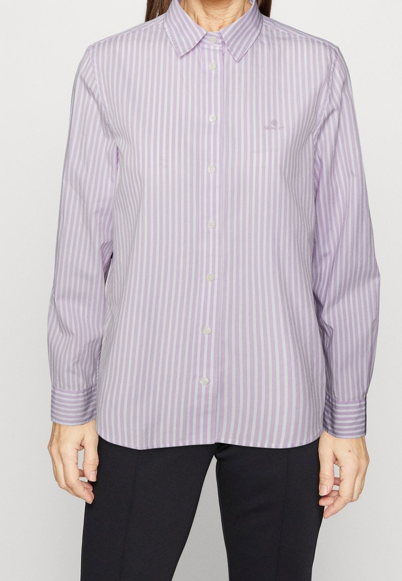 Femme portant une chemise à manches longues boutonnée avec des rayures verticales violet clair et blanc, assortie à un pantalon noir.