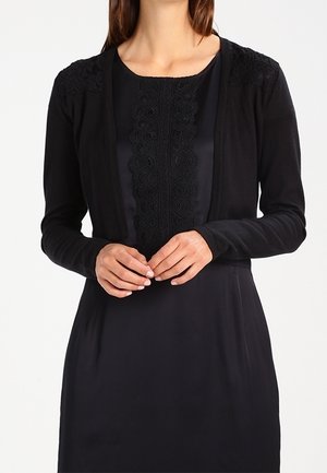 Black cardigan with lace detailing on shoulders and a textured trim at the neckline, worn over a black dress. Smooth fabric and fitted design.