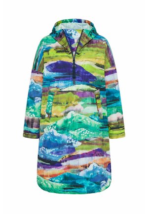 Vrouw met lang krullend haar, gekleed in een kleurrijke jas met abstract patroon, blauwe spijkerbroek en lichtpaarse sneakers, staand met gekruiste armen.