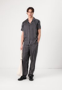 Camicia a righe navy e crema con maniche corte abbinata a pantaloni coordinati; tessuto leggero; vestibilità comoda; modello che tiene una borsa tote beige.