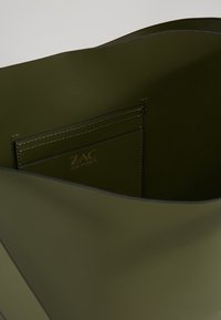 Vnitřní pohled na olivově zelenou koženou tašku, která ukazuje vyšívanou kapsu s logo ZAC Zac Posen vypáleným uvnitř.