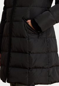Črna napihnjena bunda z quilted segmenti, ki ima zadrgo stransko žep in rebraste detajle na manšetah. Gladka tekstura, lahkoten material.