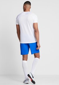 Camiseta deportiva blanca, pantalones cortos azules, calcetines blancos hasta la rodilla y zapatillas deportivas negras con detalles rojos. Tela lisa, diseño ajustado, detalles mínimos.