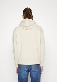 Beige Kapuzenpullover mit lässiger Passform, ausgestattet mit einer auffälligen Kapuze, gerippten Bündchen und Saum, kombiniert mit blauen Jeans.