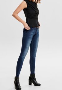 Top nero aderente con design drappeggiato, abbinato a jeans skinny blu scuro e stivaletti neri con tacco spesso, su uno sfondo neutro.