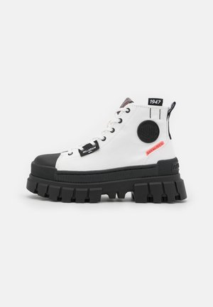 Witte hoge schoenen met zwarte rubberen neuskap en dikke zool. Voorzien van een ronde zwarte logopatch, vetergaatjes en rode accentstiksels.