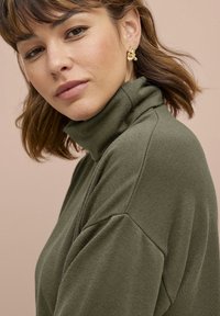 Donkergroene turtleneck trui met geribbelde textuur en verlaagde schouders, gecombineerd met gouden oorbellen, tegen een lichtroze achtergrond.