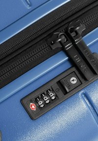 Valise rigide bleue avec une fermeture éclair noire et un cadenas à combinaison à trois roues. Texture lisse avec réflexion de la lumière sur la surface.