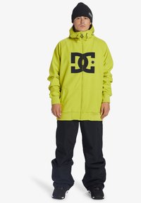 Neon grön hoodie med svart DC-logotyp, dragkedja framtill, justerbar huva, tillsammans med svarta snöbyxor och svarta skor, med en avslappnad passform.