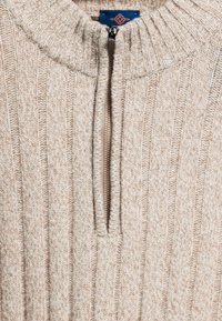 Beige gebreide trui met een ribbelstructuur en een halve ritssluiting. Opvallende details zijn de contrasterende rits en verticale strepen.