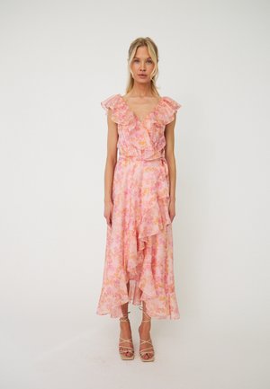 Femme blonde debout portant une robe portefeuille fleurie rose sans manches avec des volants et des sandales à talons hauts beige à brides sur fond blanc.