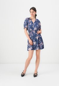 Femme se tenant dans une robe courte bleue à motifs floraux avec des manches bouffantes et des chaussures noires à talons pointus, sur un fond blanc uni.