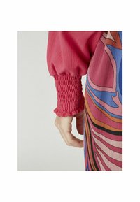 Rosa, volangärm med texturerad smock, i kombination med ett färgglatt, abstrakt mönstrat plagg med vågiga linjer i blått och orange.