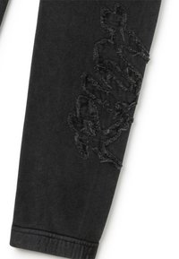 Schwarze Sweatpants mit ausgefransten Details in einem abstrakten Muster entlang eines Beins. Weiches Material mit einer enganliegenden Passform und gerippten Knöchelbündchen.