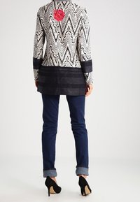 Mönstrad kavaj med svart och vit zigzag-design, prydd med en rosa blomma som accent. I kombination med mörkblå jeans och svarta högklackade skor.