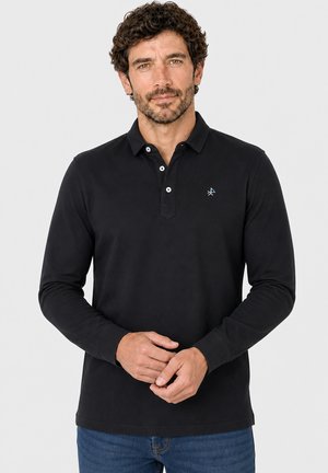 Hombre con cabello rizado oscuro y barba, vestido con una camiseta polo negra de manga larga y vaqueros azules, de pie contra un fondo claro.