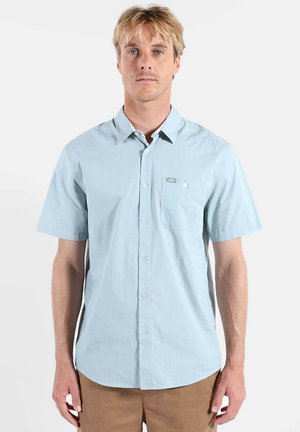 Chemise bleu clair à manches courtes en tissu doux, avec un col boutonné, une poche avant et des boutons blancs sur une texture lisse.