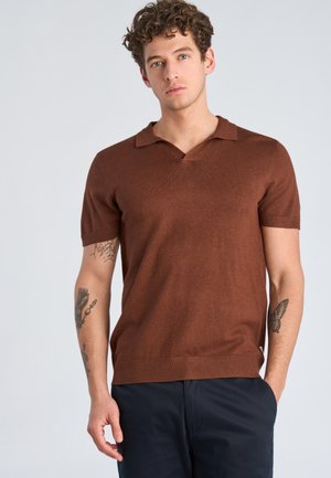 Młody mężczyzna z kręconymi włosami, ubrany w brązowy krótki sweter polo z krótkim rękawem i ciemne spodnie, pokazujący tatuaże na obu przedramionach.