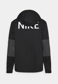 Black hooded jacket with grey elbow patches and large white NIKE text on upper back, displayed on plain light background.