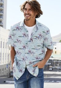 Camicia a maniche corte con bottoni caratterizzata da uno sfondo azzurro chiaro con motivi di palme rosa e onde oceaniche. Indossata sopra una t-shirt bianca.