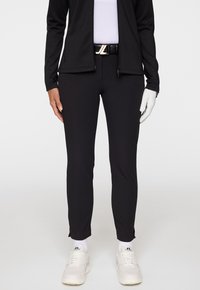 Person som bär svarta slim fit-byxor, vita sneakers med logotyp, svart bälte med guldkåpa, svart jacka, vit handske på höger hand och vit skjorta.