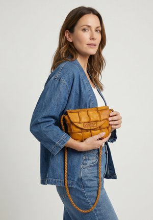 Frau in Jeansjacke und Jeans hält eine senfgelbe, gesteppte Leder-Umhängetasche mit "VENEZIA" auf der Klappe.