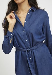 Žena v modré džínsové šate, které mají knoflíky a vázaný pás, s vrstevnatými zlatými náhrdelníky, upravuje si límec jednou rukou.