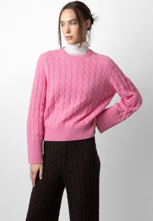 Donna che indossa un maglione rosa a trecce sopra un dolcevita bianco, con pantaloni neri dalla trama, posando davanti a uno sfondo chiaro e uniforme.