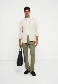 Lys beige skjorte med knapper, hvid undertrøje, olivengrønne bukser, brune loafers og en sort tote bag. Simpel design, neutrale farver.