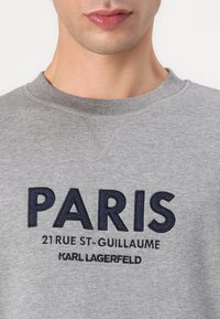 Felpa grigia con testo ricamato blu navy "PARIS", con un testo più piccolo sottostante che dettaglia un indirizzo e il nome del marchio. Texture del tessuto morbida.