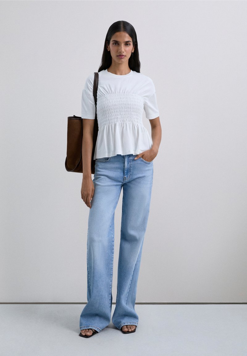 White peplum top with smocked detailing, paired with light blue wide-leg jeans. Brown shoulder bag, black open-toe sandals, and straight hair.
