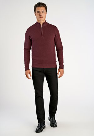 Bordeaux gebreide pullover met een halflange rits kraag, geribbelde manchetten en onderrand, gecombineerd met zwarte slim-fit broeken en zwarte schoenen, op een effen achtergrond.