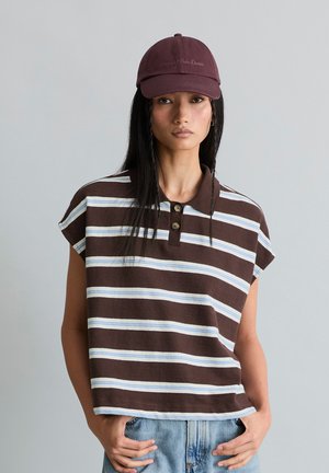 Jeune femme aux longs cheveux noirs portant un polo sans manches rayé marron, un jean bleu clair et une casquette de baseball marron foncé.