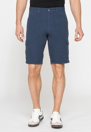 Uomo che indossa pantaloncini cargo blu navy e sneakers bianche, in piedi davanti a uno sfondo chiaro e semplice.